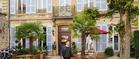 Un patio con mobiliario exterior, un árbol y un edificio con contraventanas.