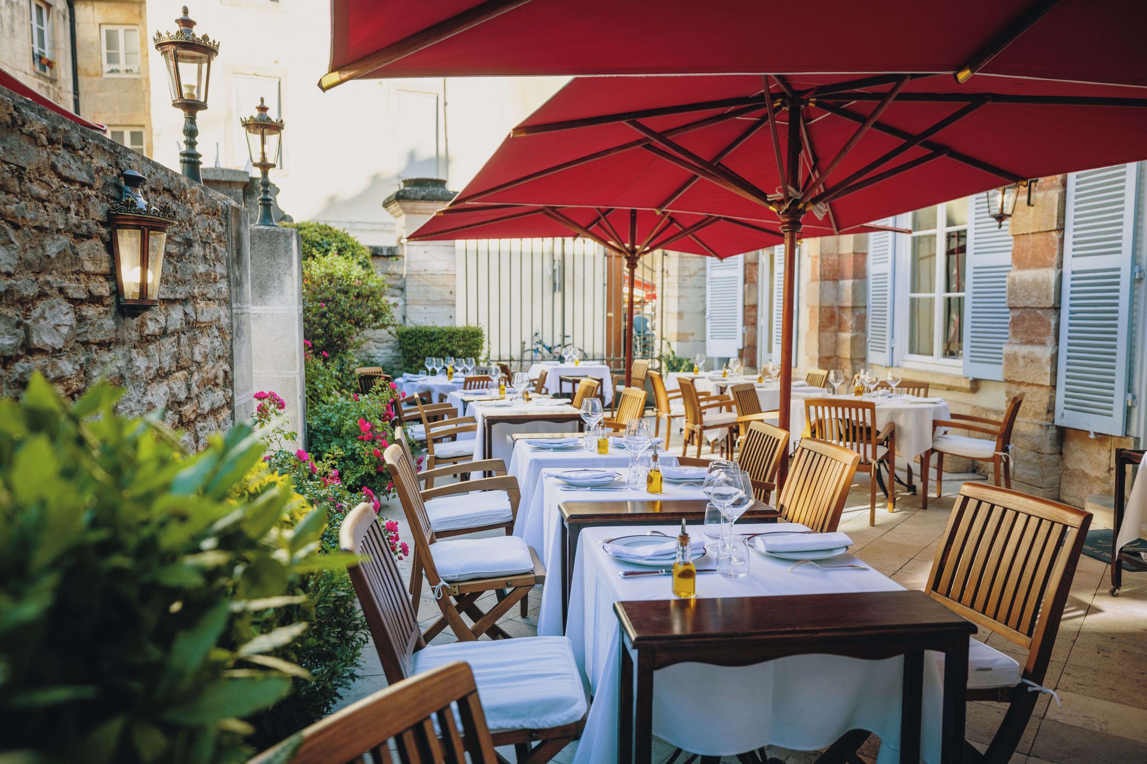 An outdoor dining area with tables and chairs, red umbrellas, and stone walls.