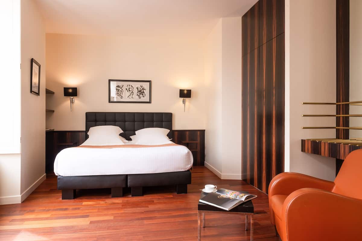 A hotel room with a large bed, two wall-mounted lamps, a framed picture, a wooden floor, and a small table with a magazine.