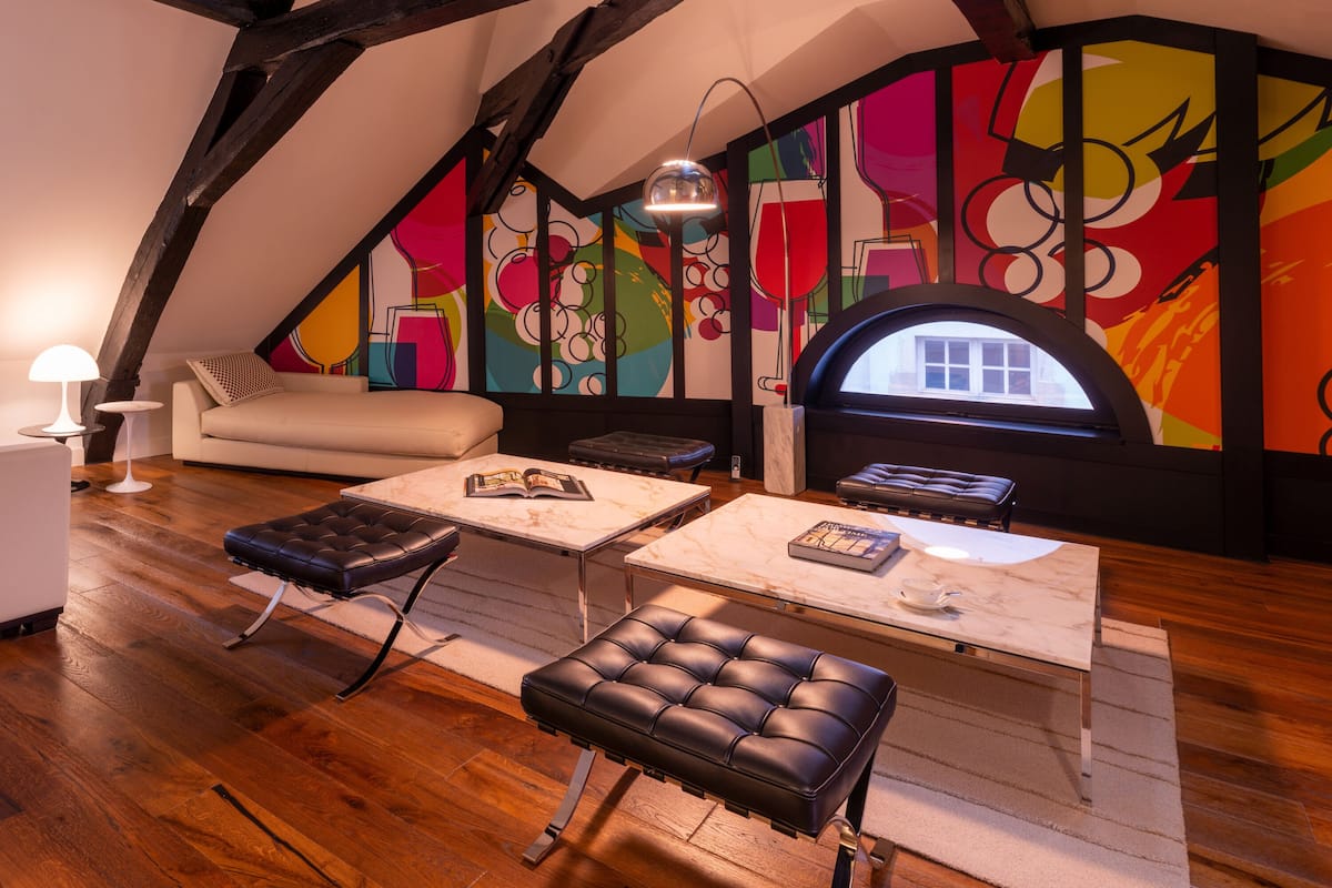A loft-style room with exposed wooden beams, a colorful abstract mural, a white sofa, a coffee table, and a leather ottoman.