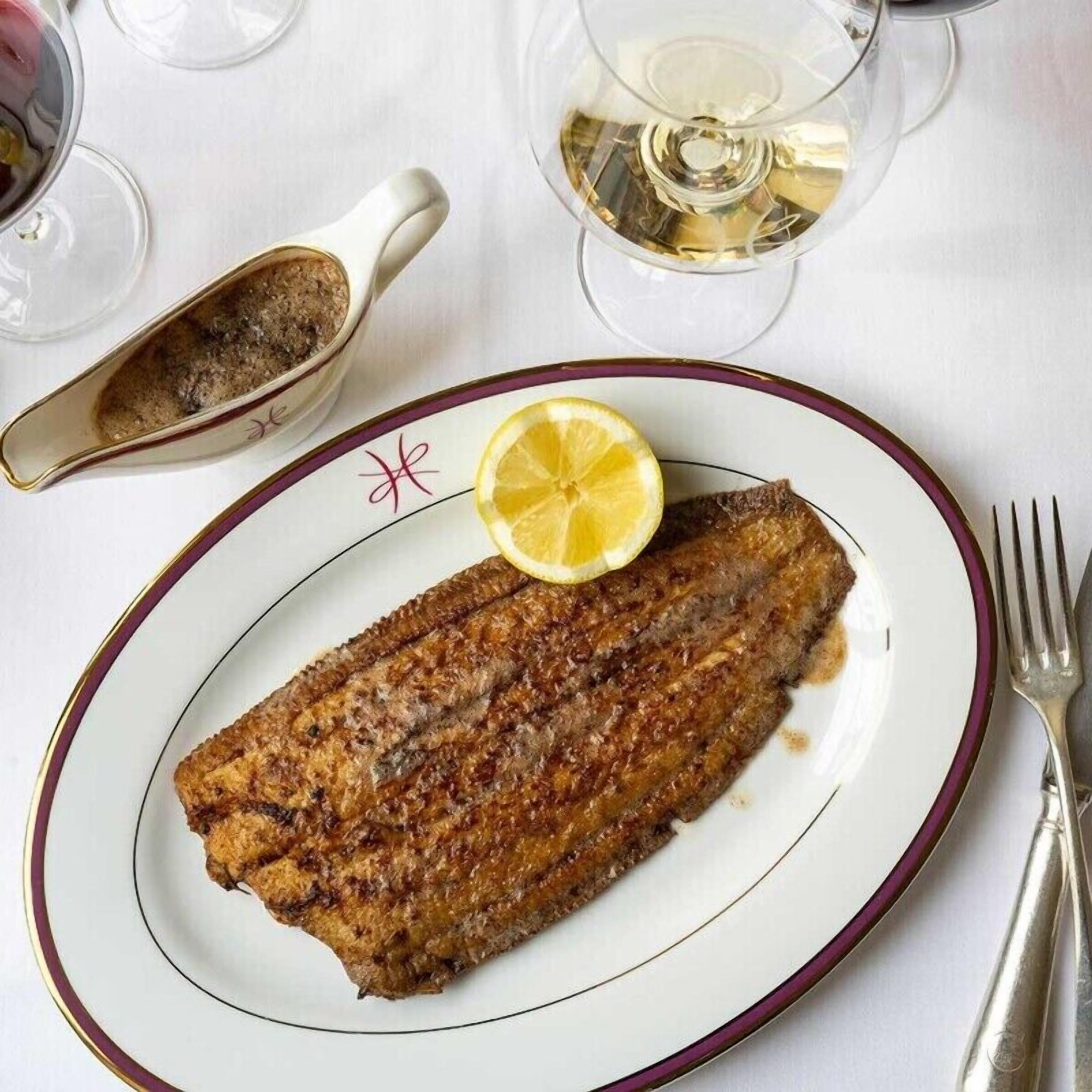 Un plato con pescado a la parrilla, una rodaja de limón y un pequeño cuenco con condimentos sobre una mesa, junto a un tenedor y una copa de vino blanco.