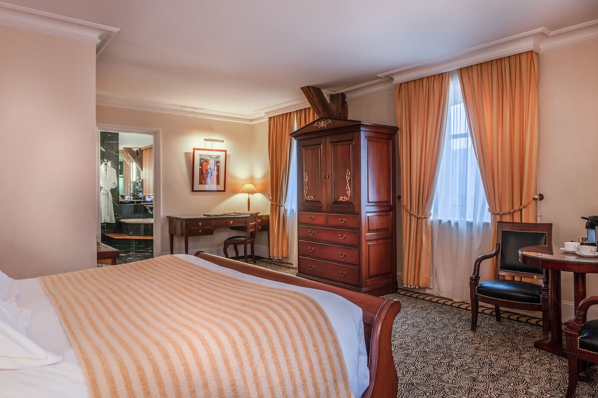 A hotel room with a large bed, a wooden wardrobe, a desk with a chair, and a small table with a chair.