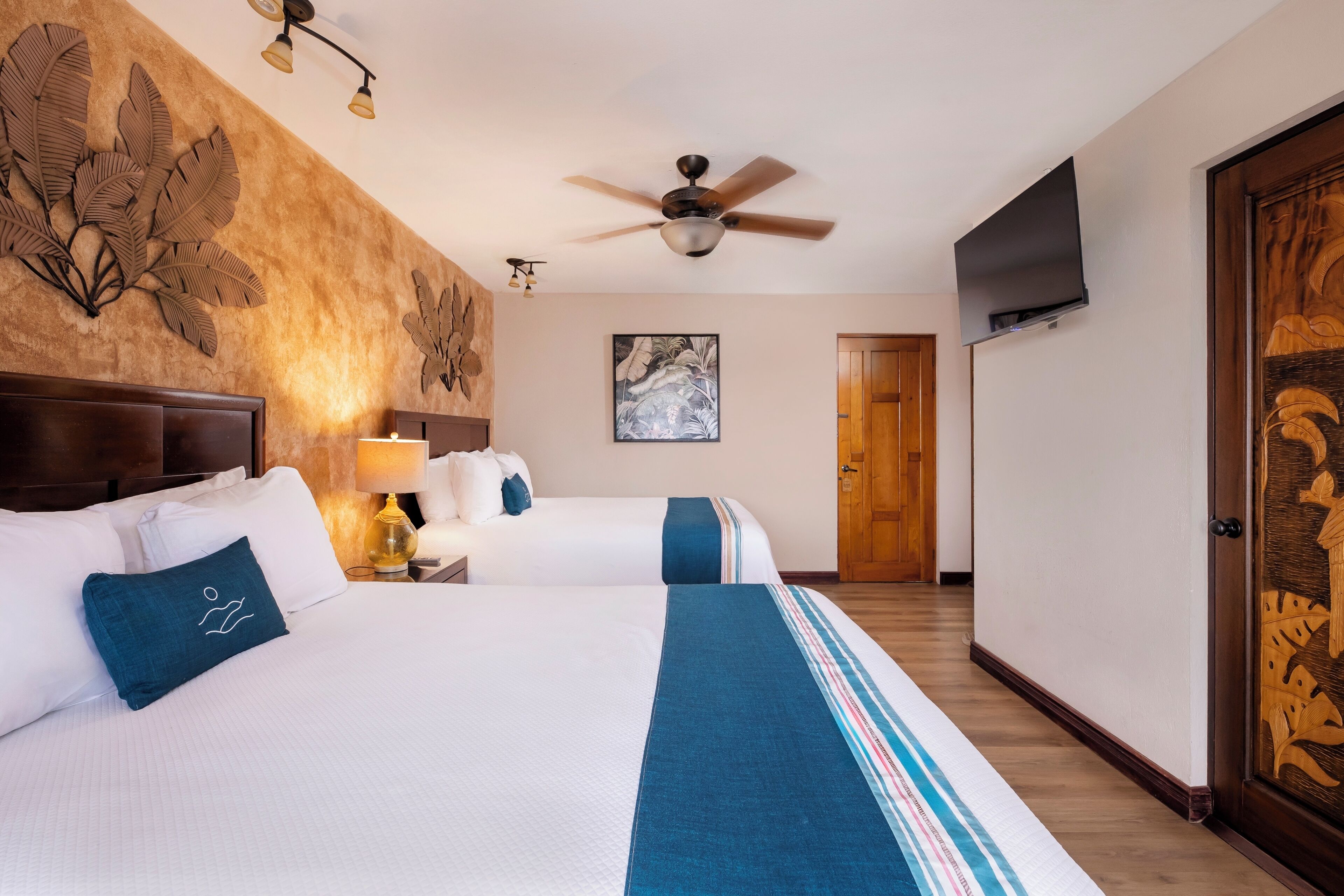 A hotel room with two beds, a ceiling fan, a painting, and a wooden door.