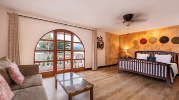A room with a wooden floor, a sofa, a bed, a table, and a large arched window.