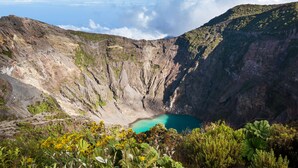 A crater with a turquoise lake surrounded by rugged terrain and vegetation.