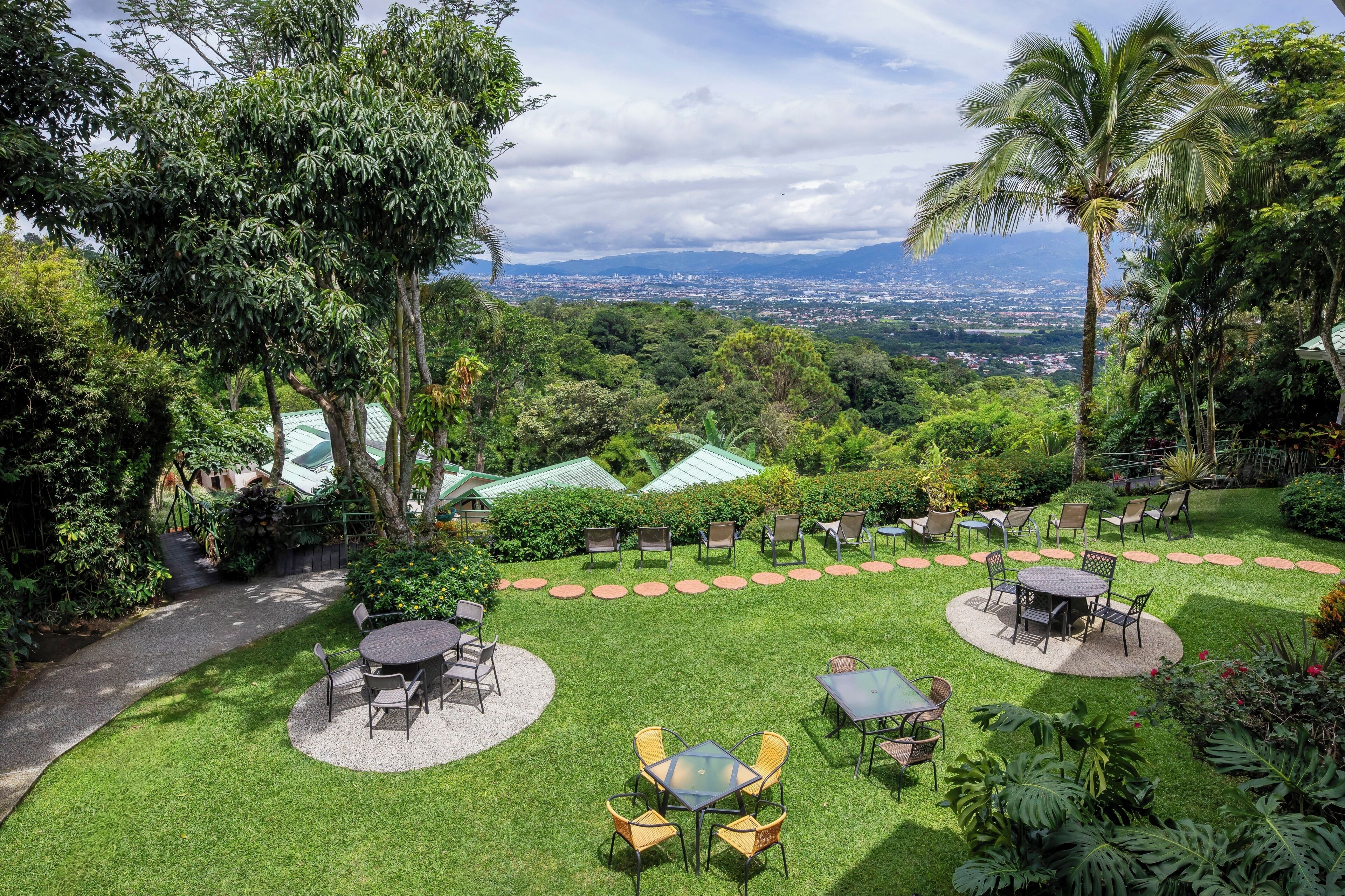 Un jardín exuberante con mesas al aire libre y una vista panorámica de la ciudad al fondo.