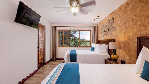 Een hotelkamer met twee bedden, een plafondventilator, een aan de muur bevestigde televisie en een raam met uitzicht op groen.