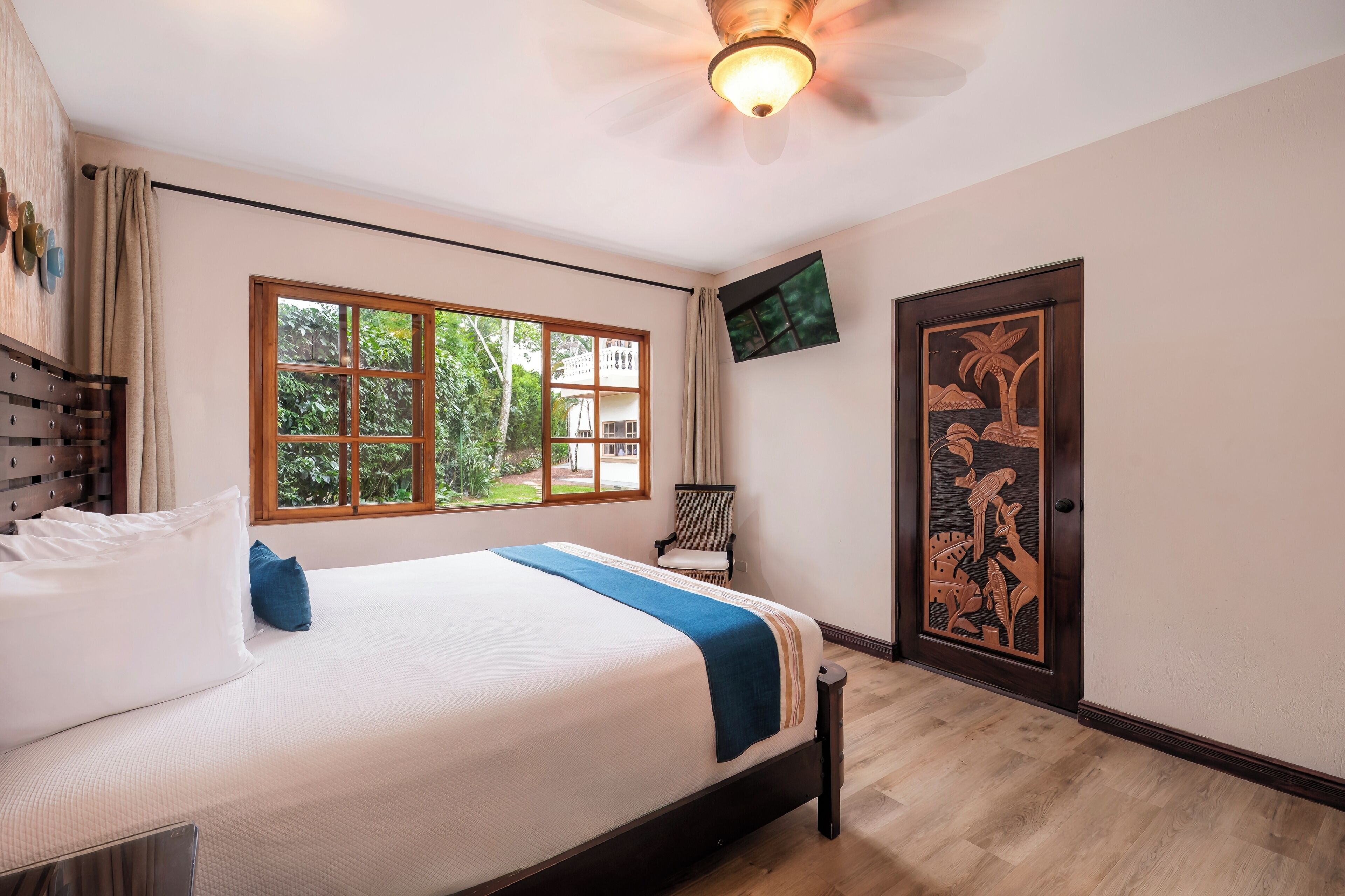 A hotel room with a bed, a window with a view of greenery, a ceiling fan, and a door with a decorative panel.