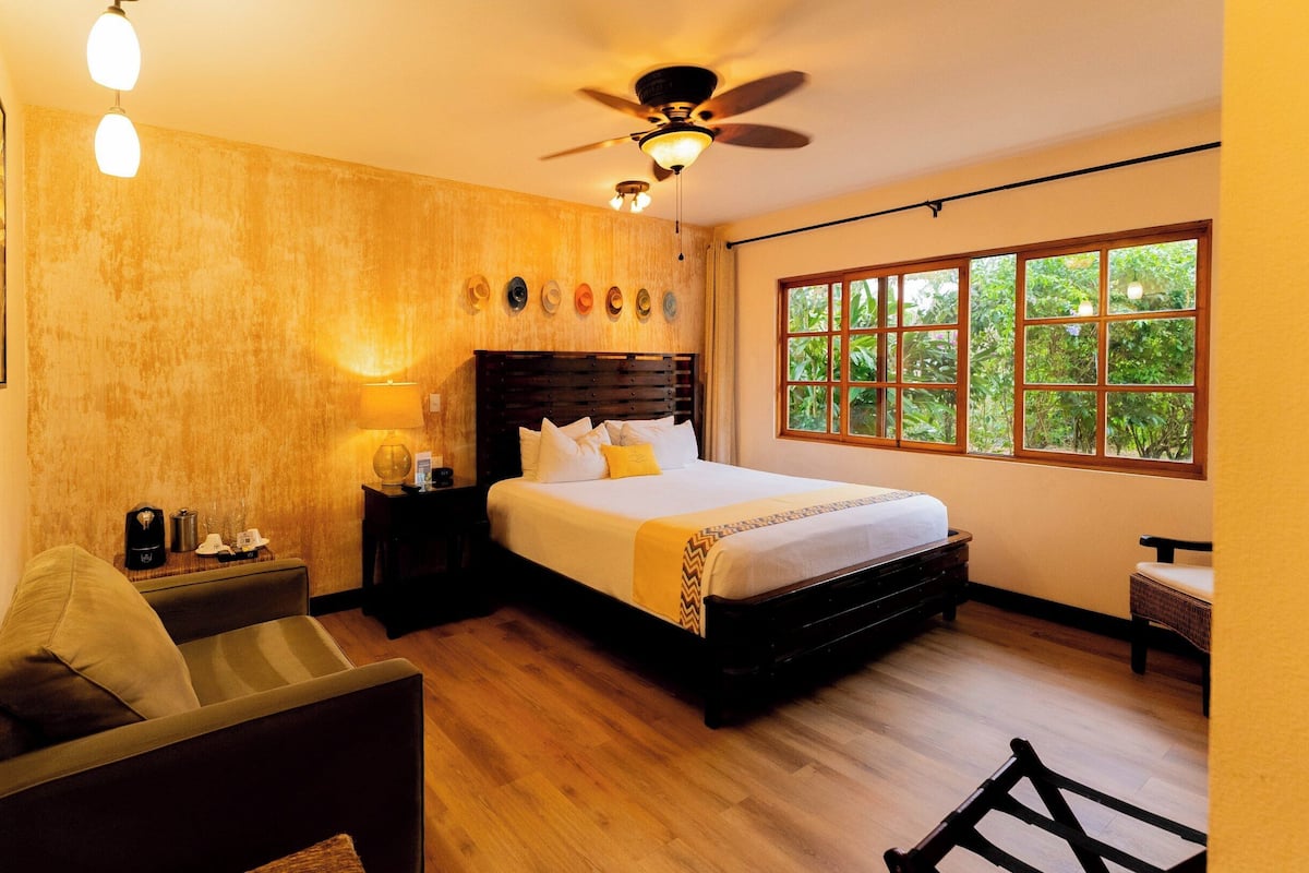 Une chambre d’hôtel avec un plancher en bois, un lit avec une literie blanche, une table de chevet avec une lampe, un ventilateur de plafond et une fenêtre donnant sur un paysage verdoyant.