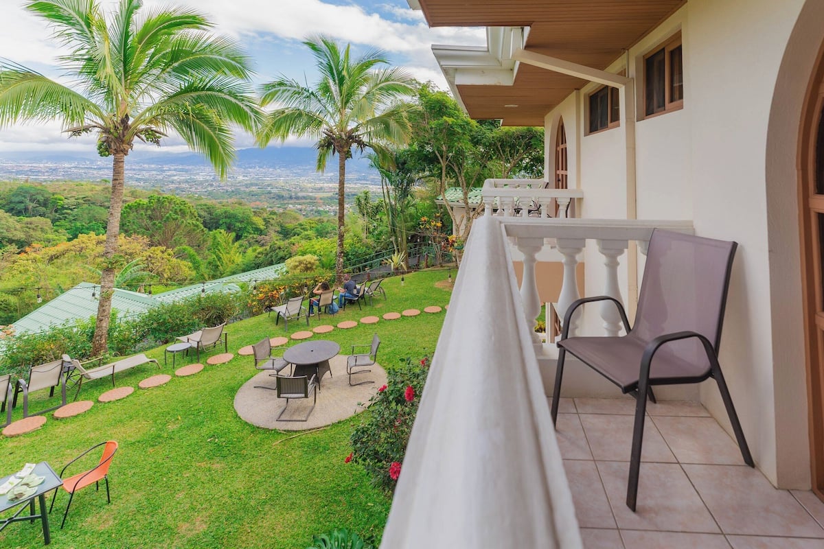 Un balcon offrant une vue sur un jardin luxuriant et sur le paysage urbain, avec un espace salon extérieur et une table.