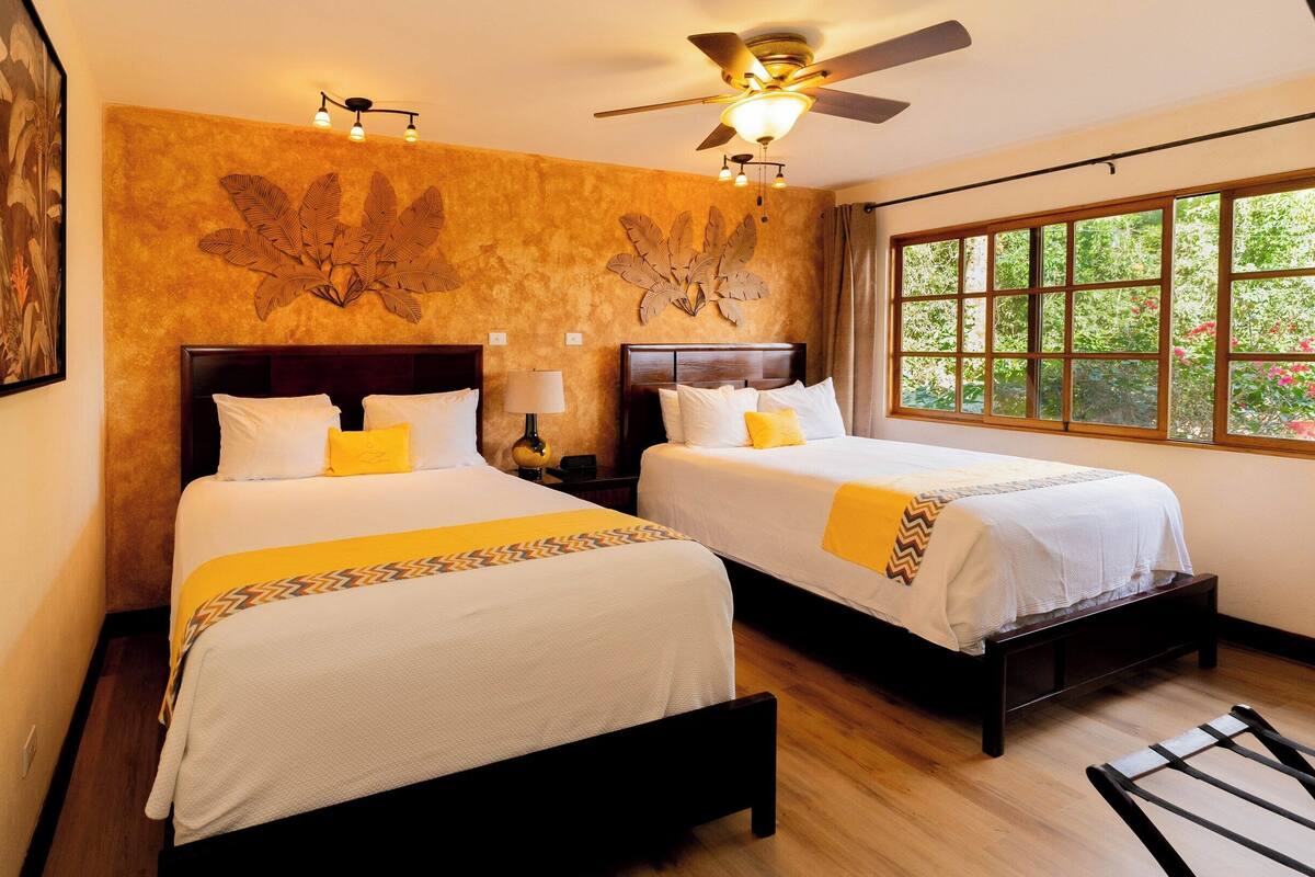 Une chambre d’hôtel avec deux lits, des têtes de lit en bois, une grande fenêtre donnant sur un paysage verdoyant, un ventilateur de plafond et des œuvres d’art accrochées au mur.