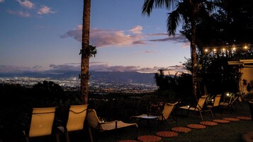 Una terraza con sillas y una mesa, con vistas a un paisaje urbano al atardecer.