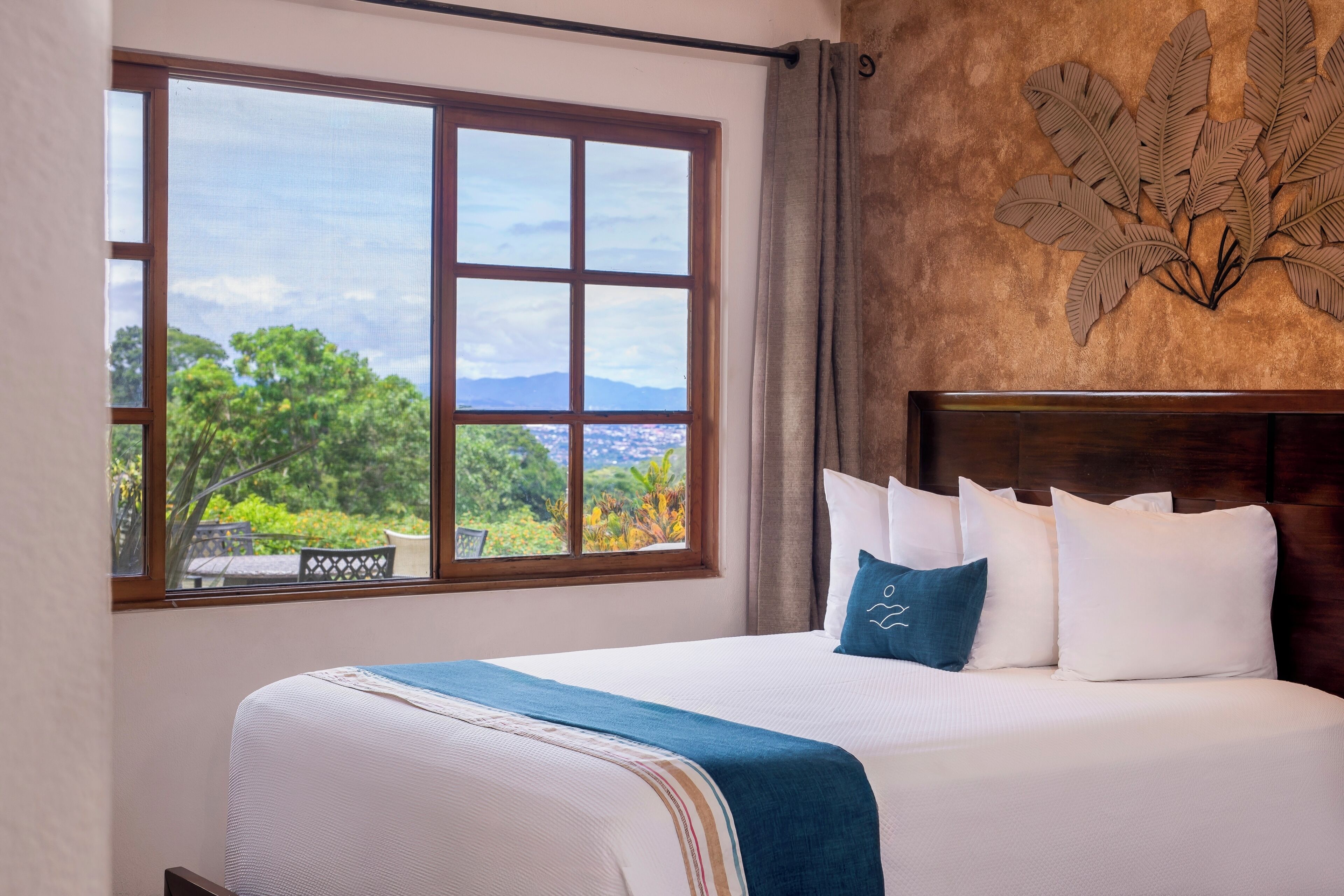 Ein Schlafzimmer mit großem Fenster, einem Bett mit weißer Bettwäsche und einer blauen Tagesdecke sowie einer Wanddekoration.
