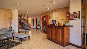 Un vestíbulo con mostrador de recepción, una escalera, una zona de estar con sofá y sillón, y una pared con un cuadro enmarcado y un cartel.