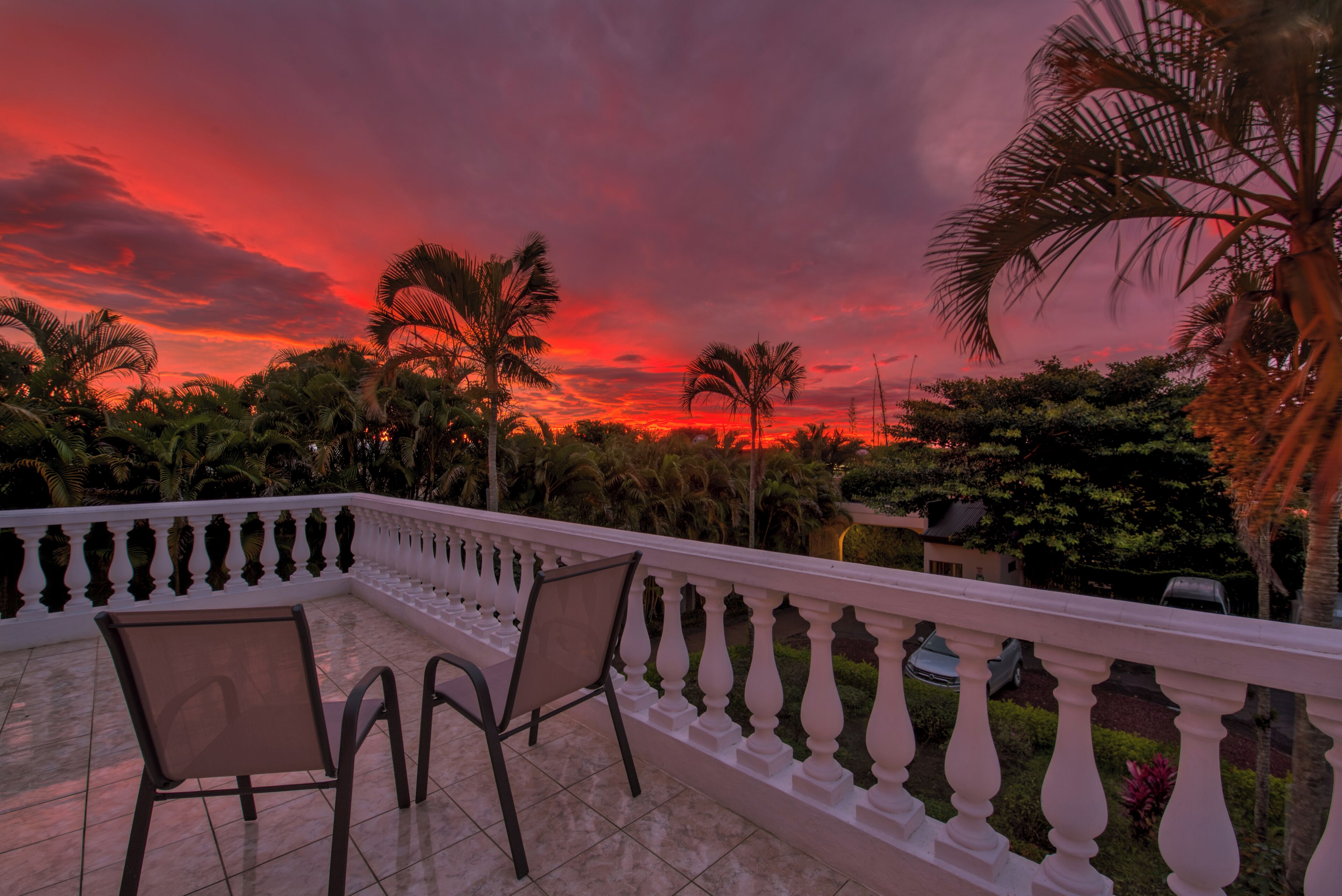 A balcony with two chairs and a railing, overlooking a garden with palm trees and a sunset.