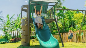 Un niño en un tobogán verde en un parque infantil, con una cerca de madera y follaje al fondo.