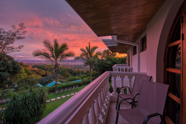Un balcón con vista a un paisaje urbano al atardecer, rodeado de follaje y palmeras.