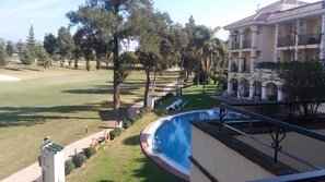 A pool area with a view of a golf course, surrounded by trees and a building with balconies. - Hotel Tamisa Golf (Mijas)