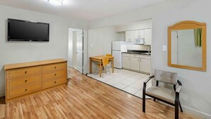 Een knus leefruimte met een houten vloer, een flatscreen televisie, een houten dressoir, een eettafel met stoelen, een keuken met witte kastjes, en een spiegel aan de muur.