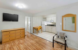 A compact living space with a wooden floor, a flat-screen TV, a wooden dresser, a dining table with chairs, a kitchen area with white cabinets, and a mirror on the wall.