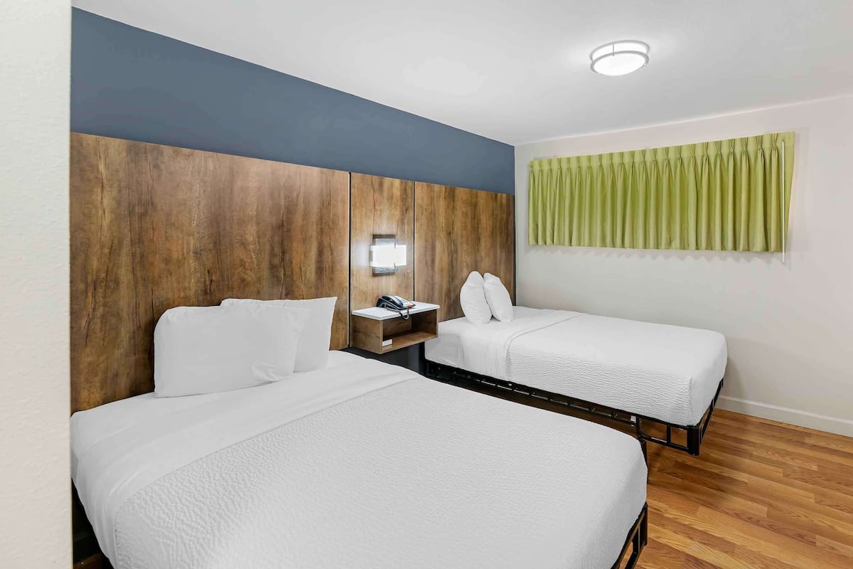 A hotel room with two beds, a wooden headboard, a bedside table with a lamp, and a green curtain.
