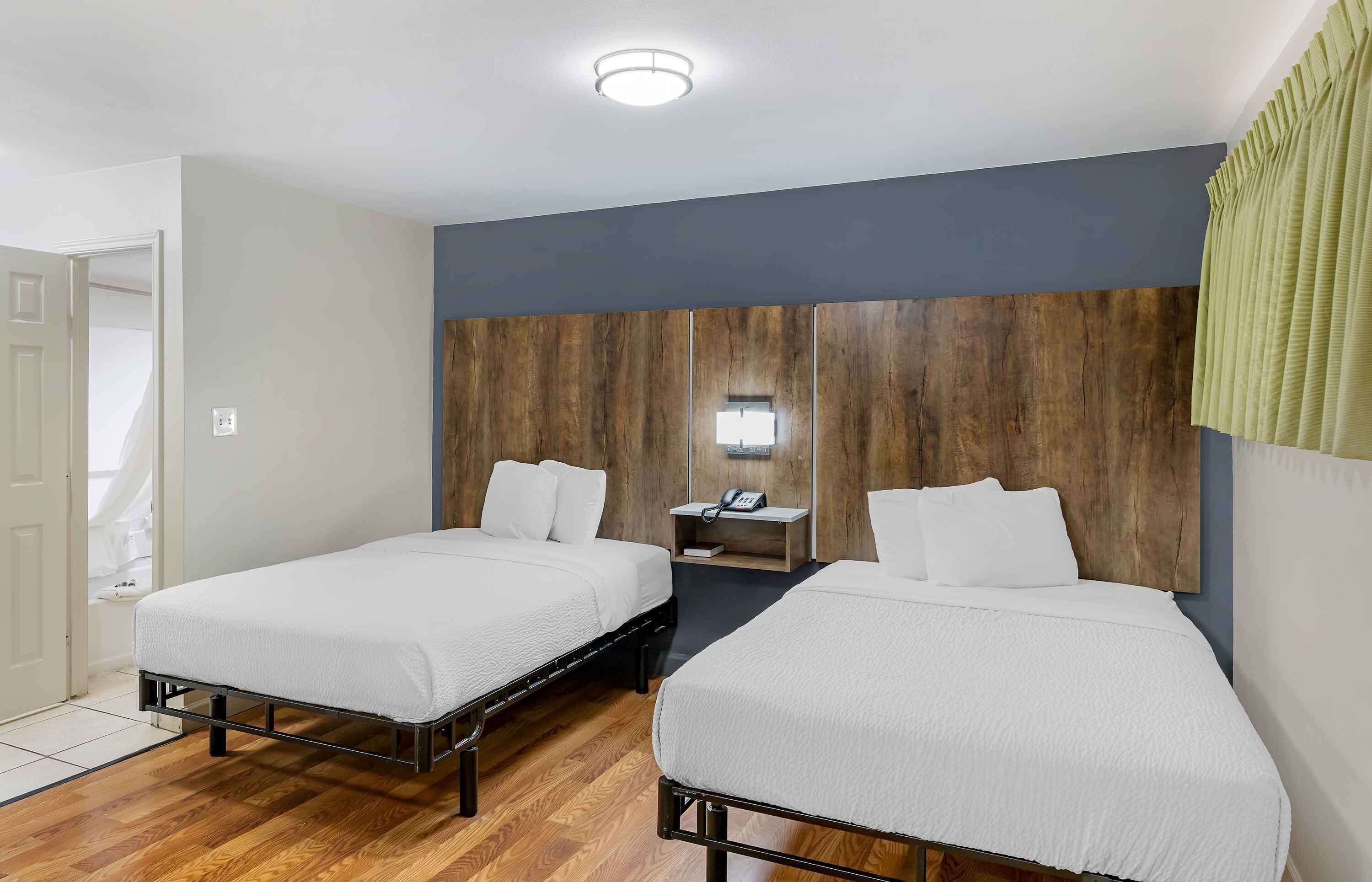 A hotel room with two single beds, a wooden headboard, a bedside table with a lamp, and a door leading to a bathroom.