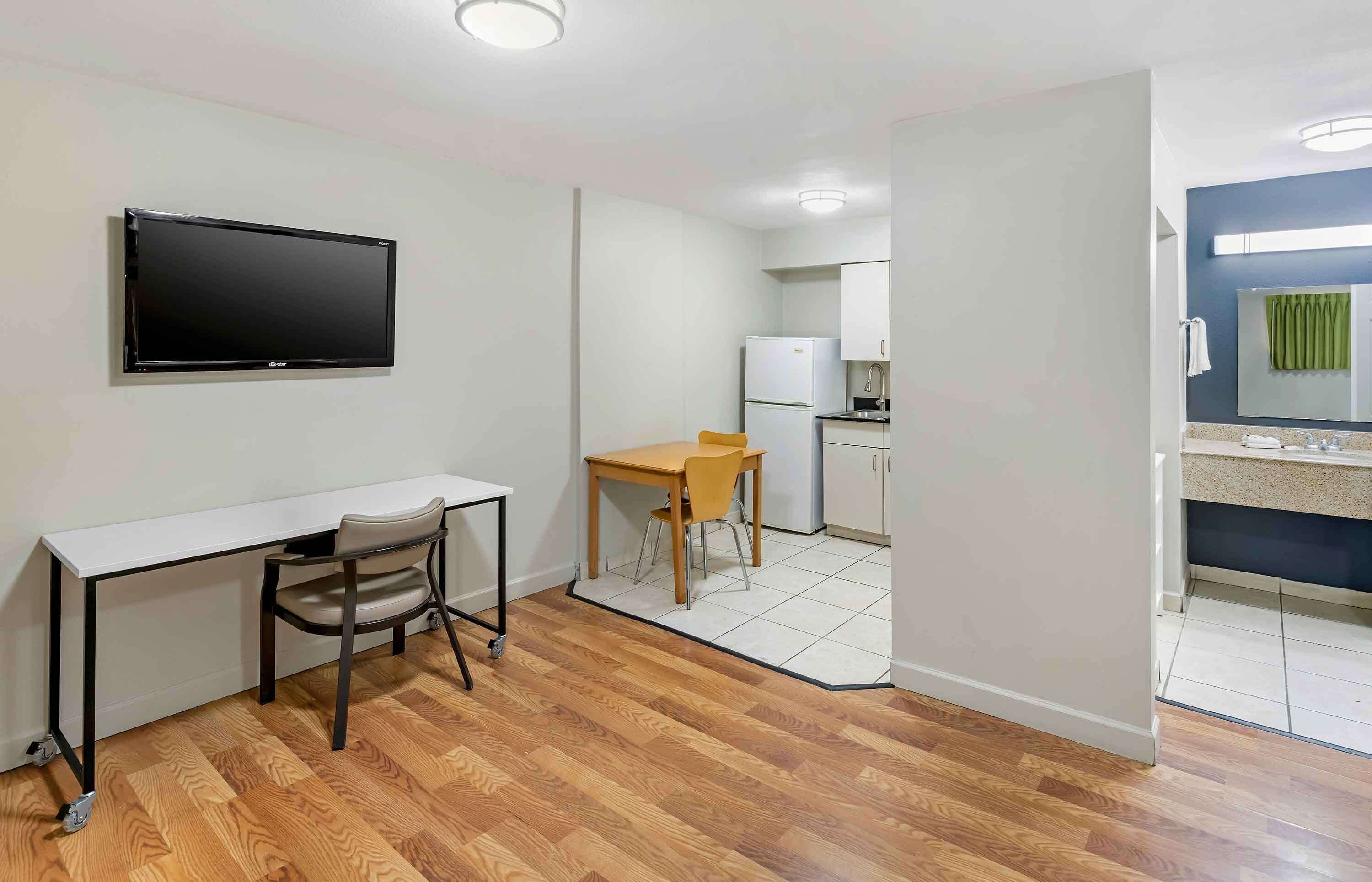 A compact room with a desk, chair, and a small table. A television is mounted on the wall. The room has a kitchenette with a refrigerator, sink, and cabinets.