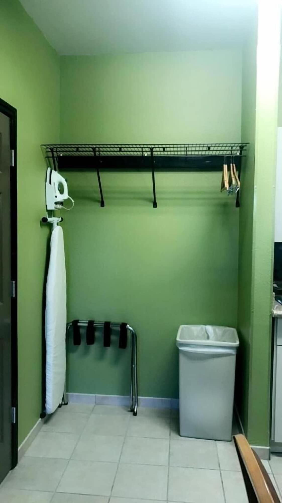 Un baño pequeño con paredes verdes, un estante de metal con productos de limpieza, una toalla blanca y un cubo de basura.