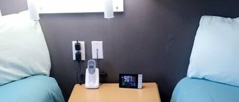A hotel room with two single beds, a bedside table with a phone and a digital clock, and a wall-mounted lamp.
