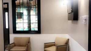 Una habitación pequeña y bien iluminada con dos sillones beiges, una mesa de centro de madera y un suelo a cuadros. Se ve una ventana con marco negro y vistas a edificios.