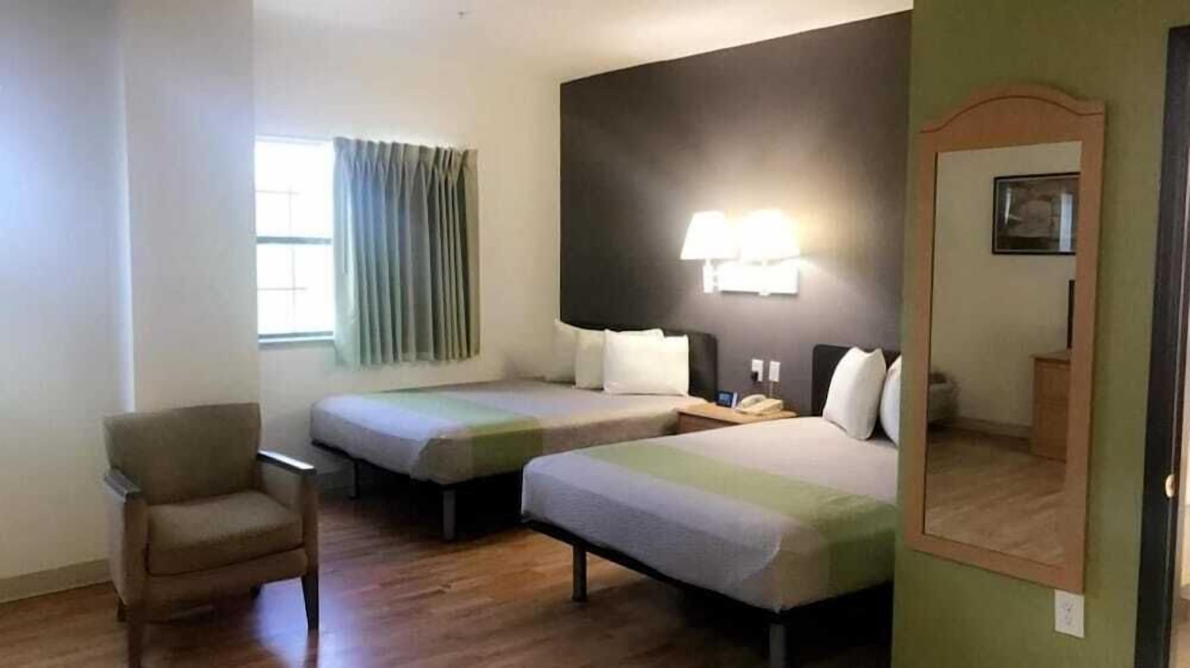 Una camera d'albergo con due letti singoli, una sedia, un comodino con lampada, uno specchio e una finestra con tende.