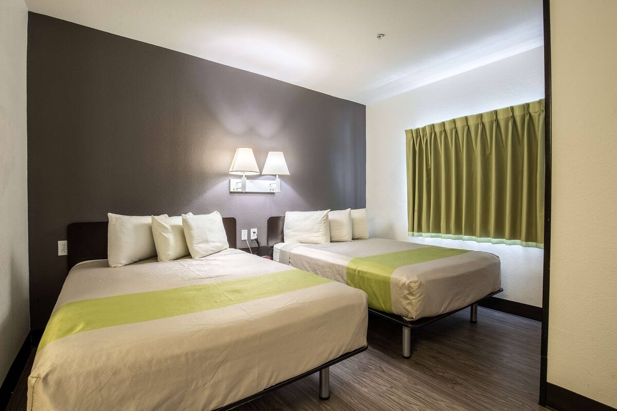 A hotel room with two beds, a nightstand, a lamp, and a window with curtains.