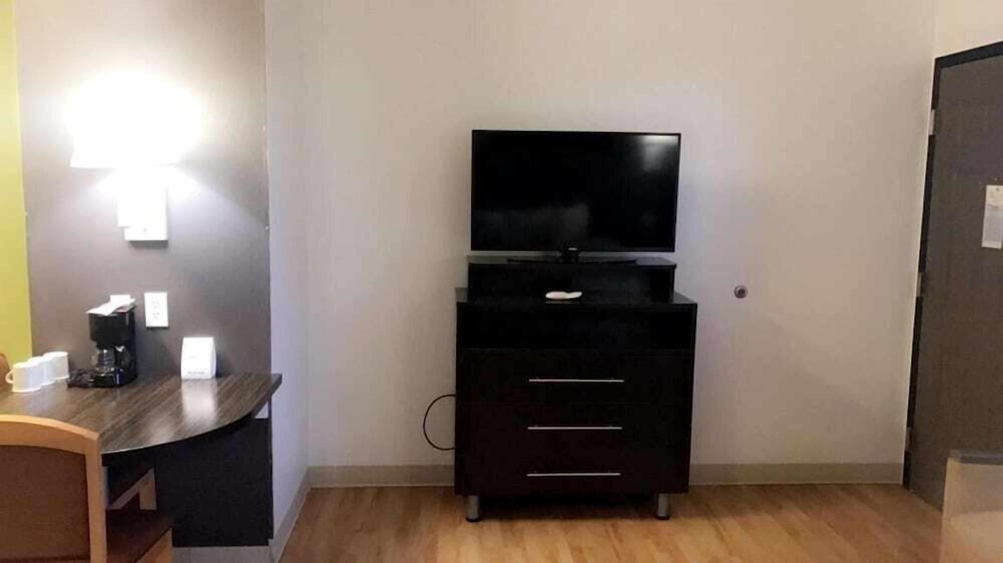 A small, modern room with a dark wooden dresser, a flat-screen TV on top, and a desk with a coffee maker and cups.