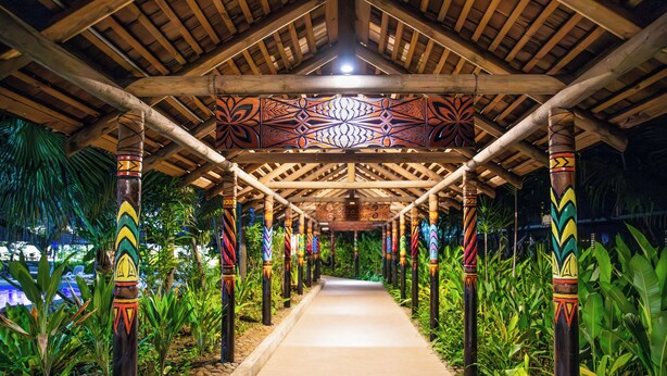 A covered walkway with intricate wooden carvings and colorful patterns on the pillars, surrounded by lush greenery. - Tanoa Tusitala Hotel (Apia)