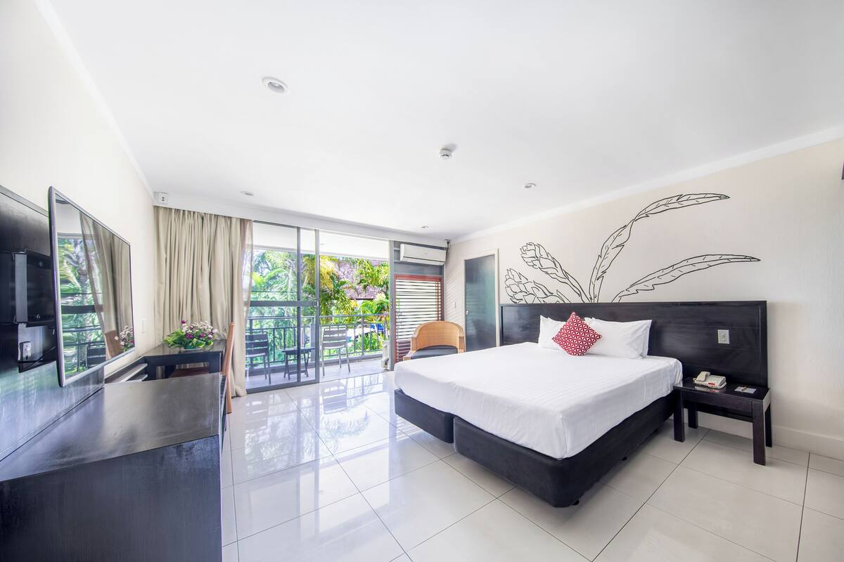 A modern hotel room with a large bed, a desk, a TV, and a balcony with a view of greenery.