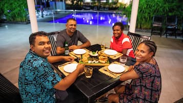 A group of people dining at an outdoor table with a pool in the background.