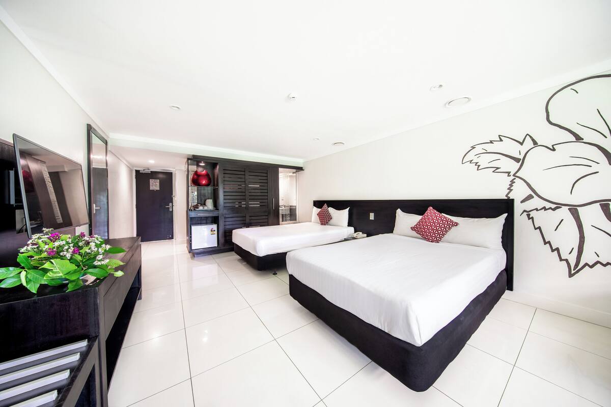 A modern hotel room with a large bed, a smaller bed, a desk with a computer, and a plant.
