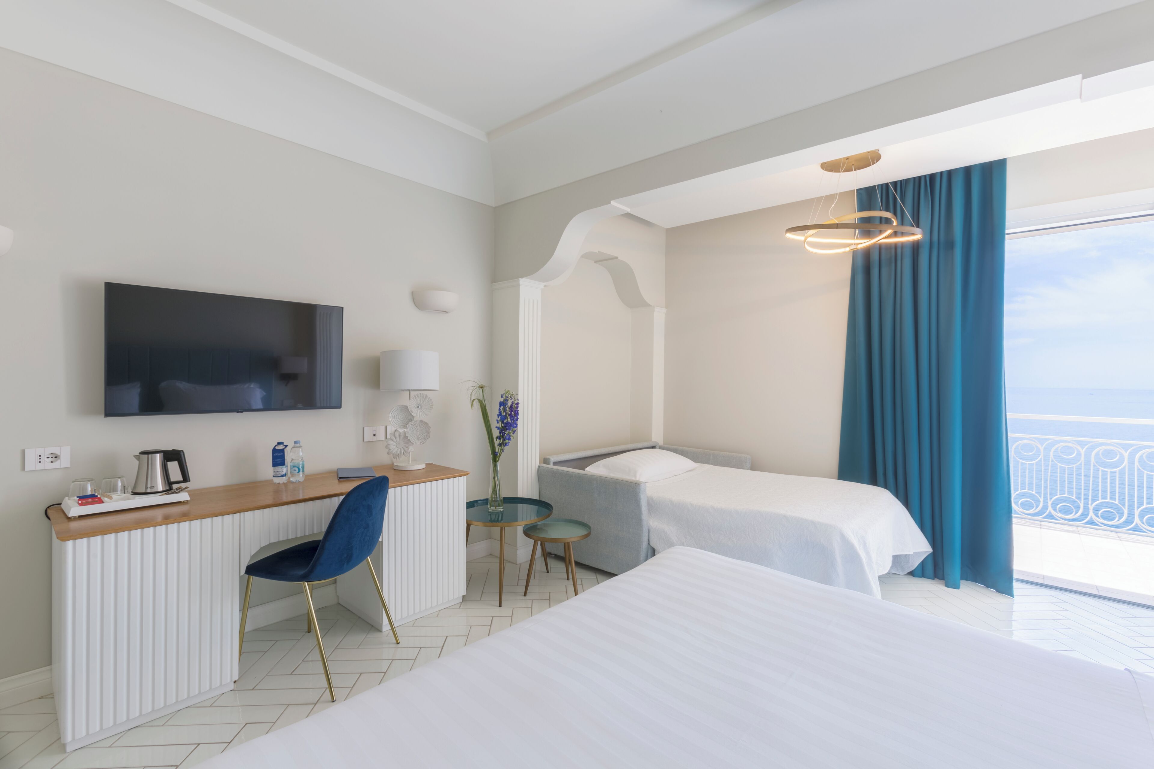 A hotel room with a bed, a desk with a chair, a TV, a small table with a vase, and a balcony with curtains.
