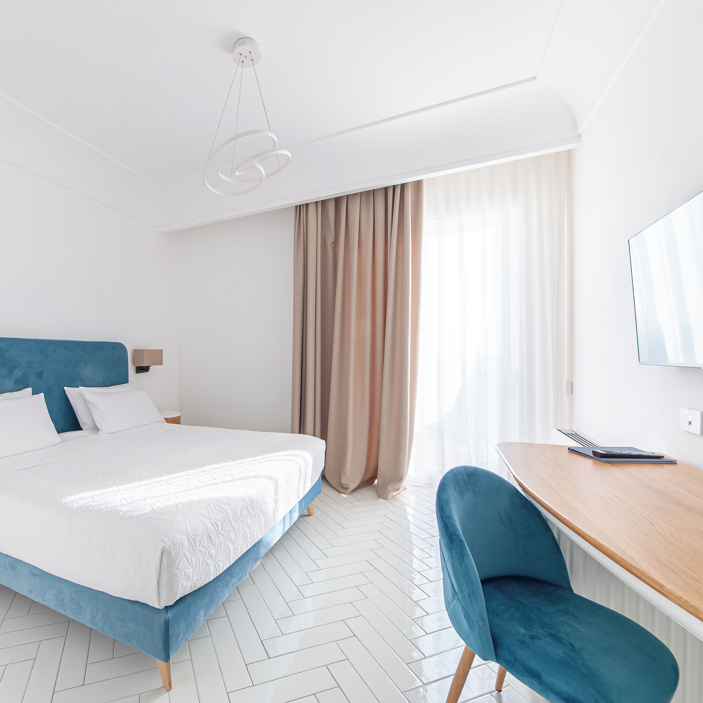 A hotel room with a bed, a desk, a chair, a window with curtains, and a ceiling light fixture.