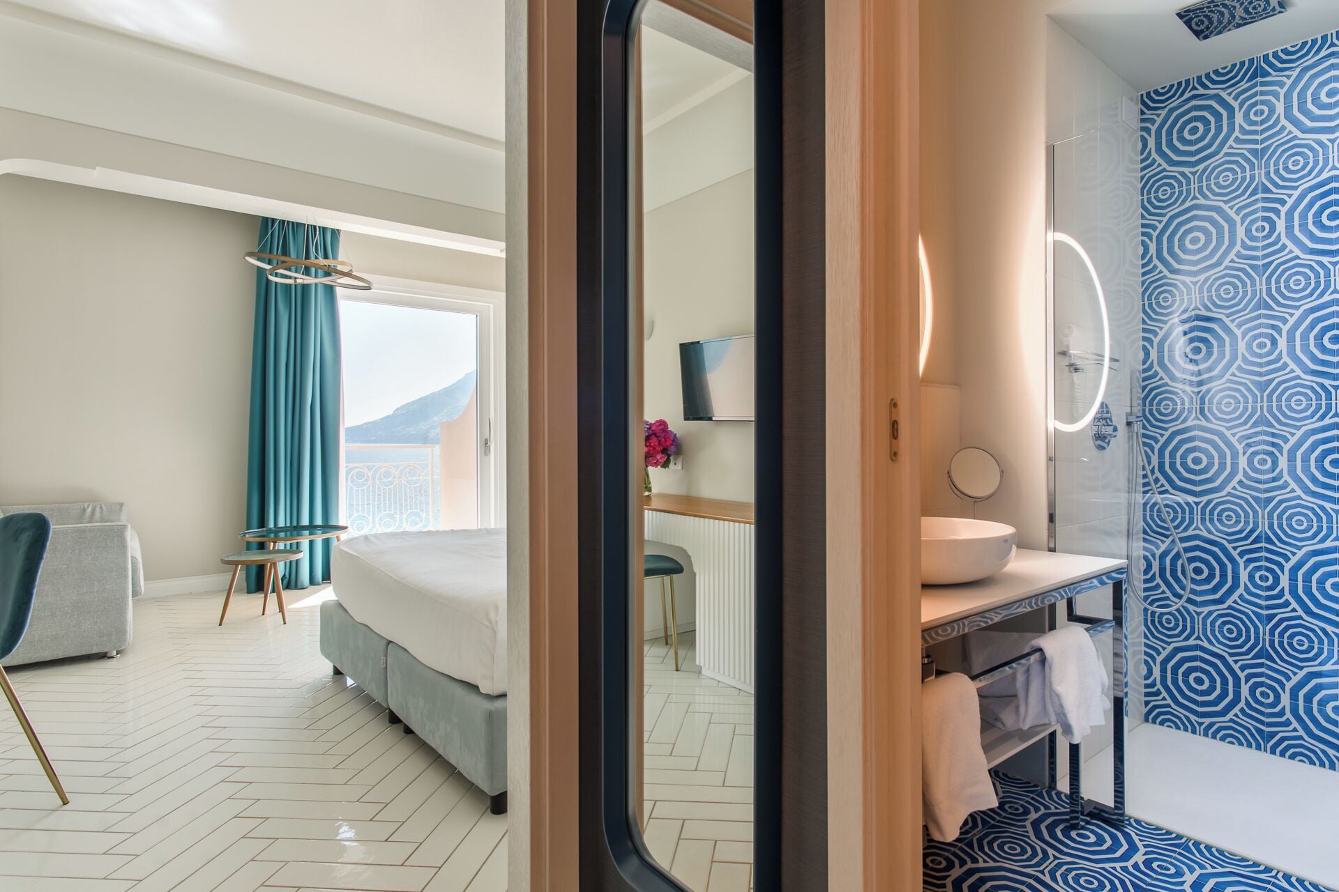 A hotel room with a bed, a desk, a bathroom with a mirror and sink, and a view of the outdoors.