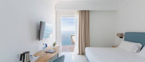 A hotel room with a bed, a desk with a kettle, a chair, a TV, and a view of the sea.