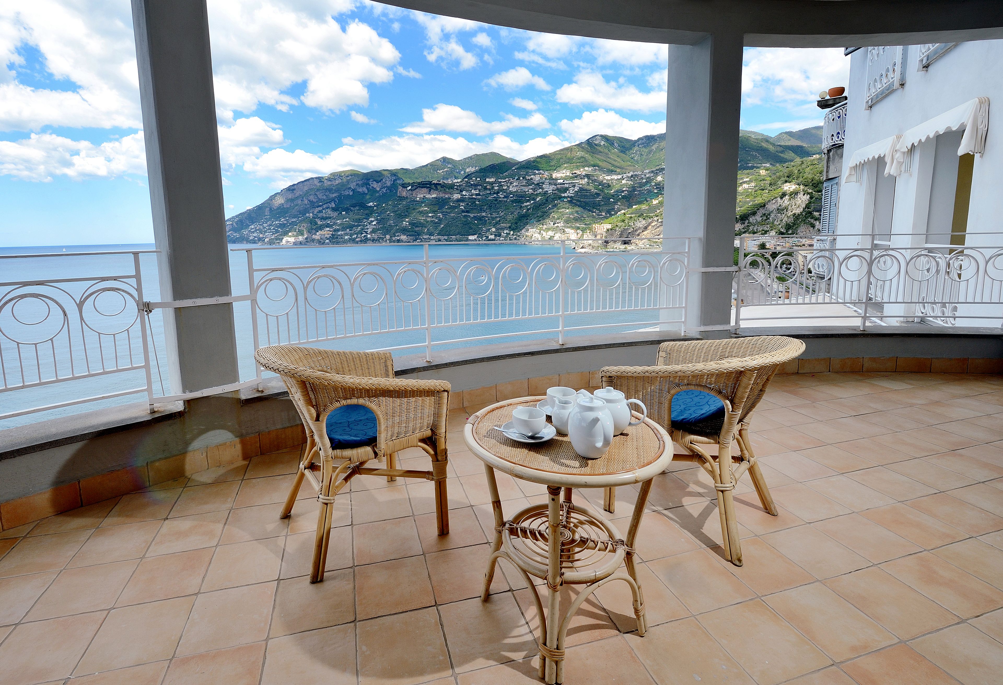 Un balcon agrémenté de fauteuils en osier et d’une petite table, offrant une vue sur un paysage côtier avec des montagnes et un ciel dégagé.