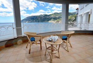 Un balcon agrémenté de fauteuils en osier et d’une petite table, offrant une vue sur un paysage côtier avec des montagnes et un ciel dégagé.