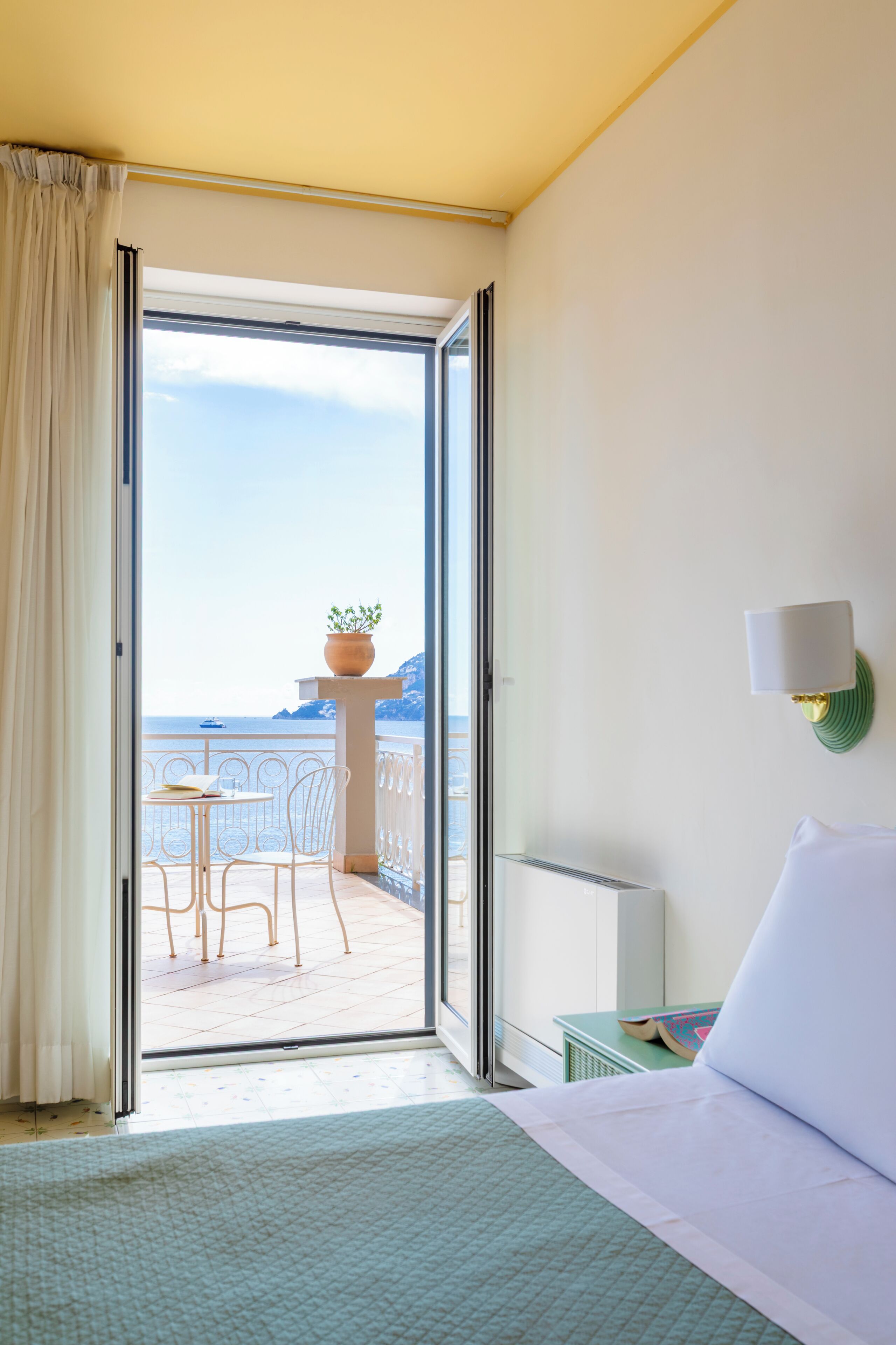 A hotel room with a bed, a nightstand, a lamp, and a view of the sea through an open door.