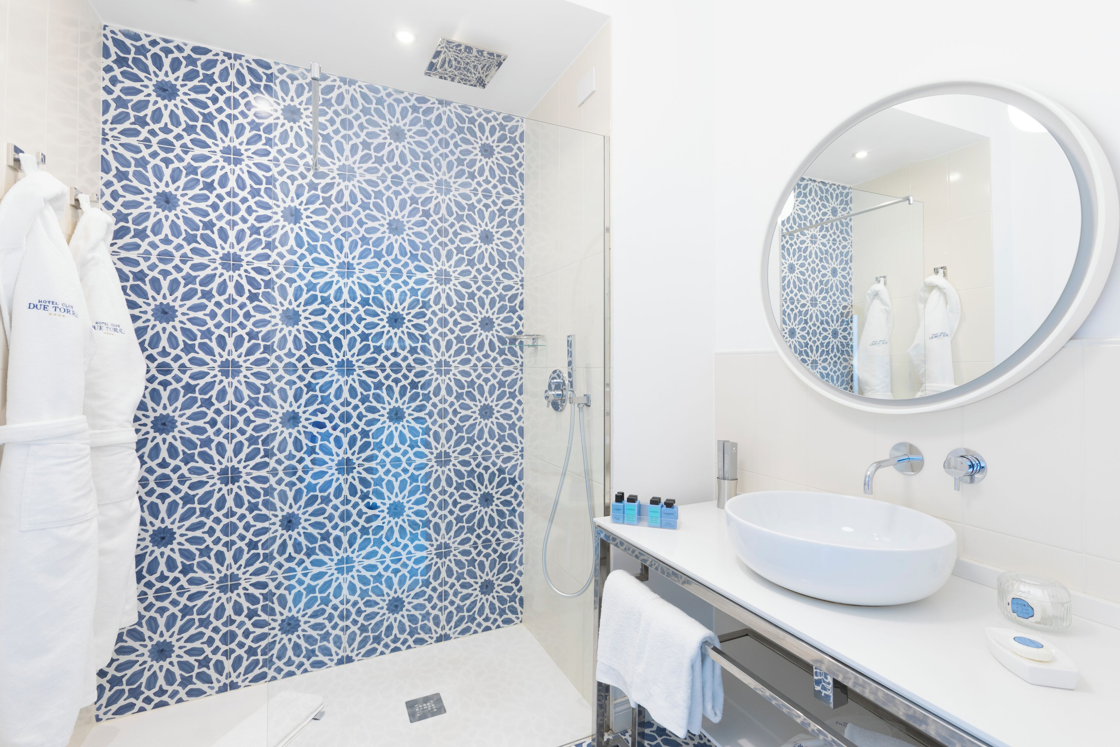 A modern bathroom with a patterned shower wall, a round mirror, a white sink, and a towel rack.