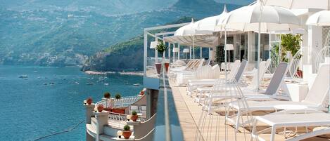A sunlit outdoor seating area with white lounge chairs and umbrellas, overlooking a body of water and a mountainous landscape.