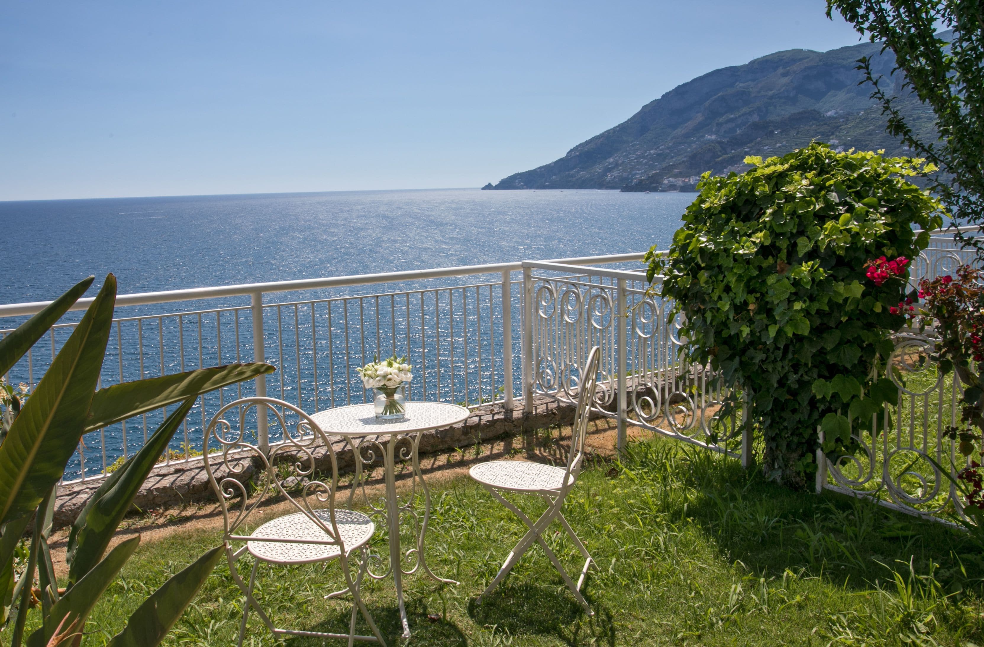 A balcony with a white metal railing, a round table with a vase of flowers, and two chairs.