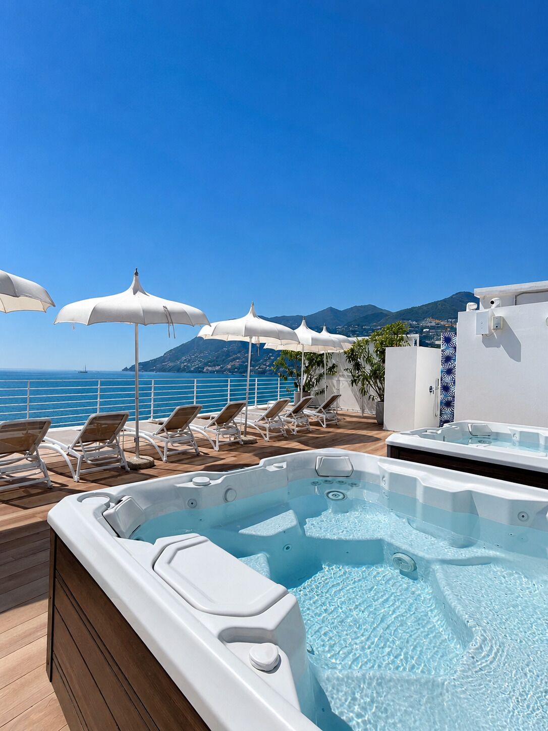 A rooftop pool area with lounge chairs and umbrellas, overlooking a body of water and hills.