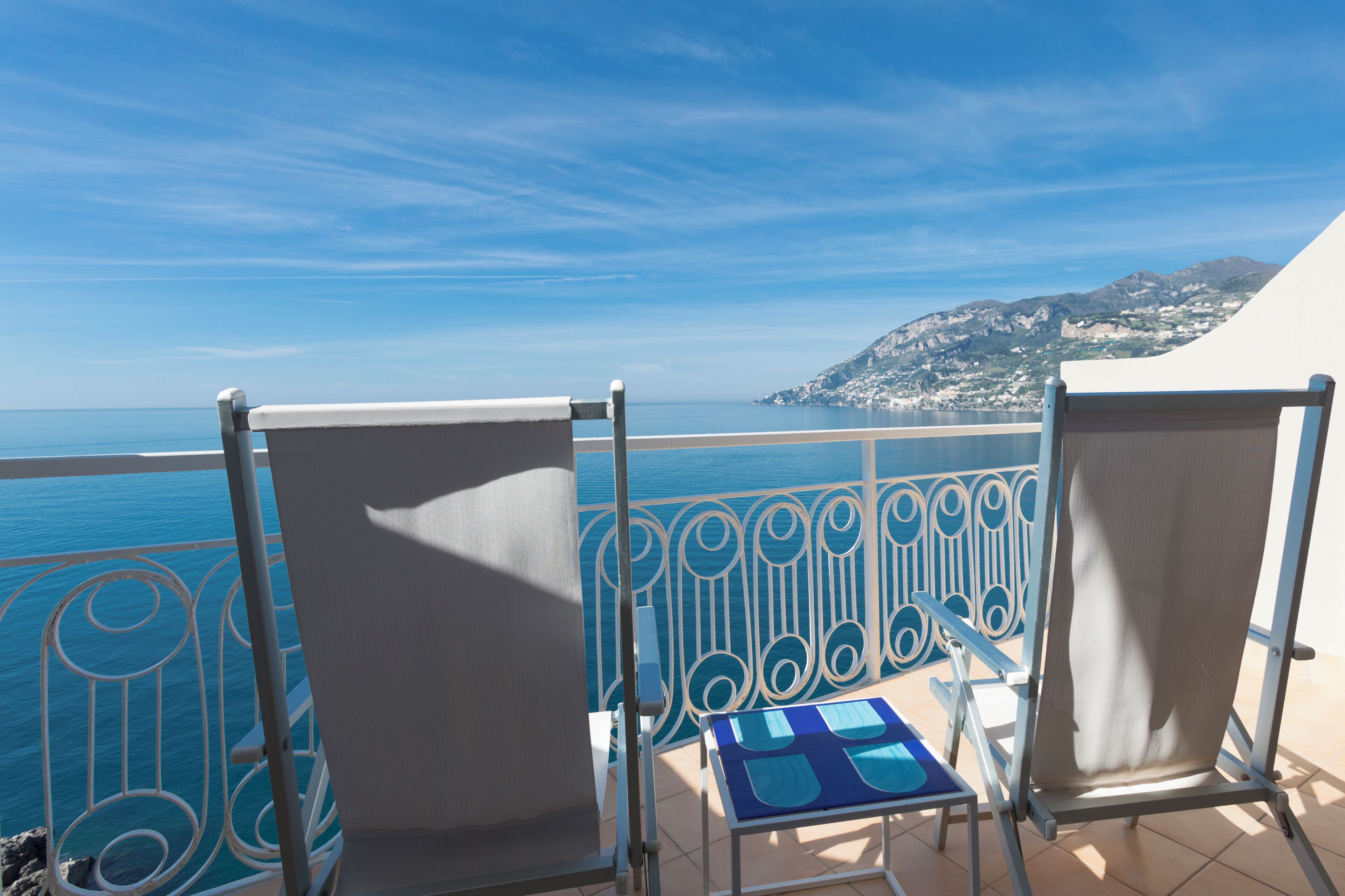 A balcony with two chairs and a small table, overlooking a coastal view with a clear sky.