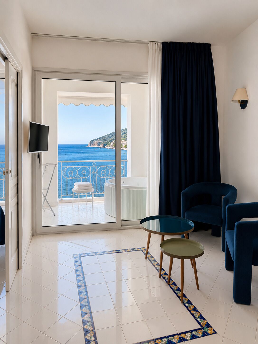 A hotel room with a balcony, a TV, a seating area with chairs, and a view of the sea.